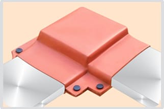A terracotta moulded shroud fitted over a right-angle busbar connection, raised into a square boss where it clears the bolted stack, with dark fastener heads visible on the flange and bright bare bars leaving it left and right.
